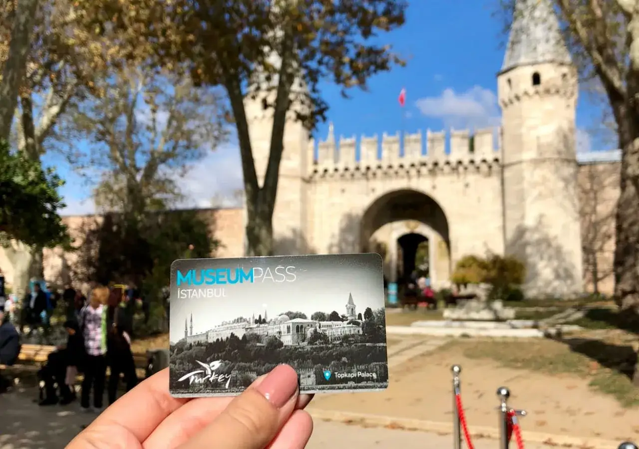 A hand holds a Museum Pass for Istanbul, Turkey, in front of the grand entrance to Topkapi Palace.