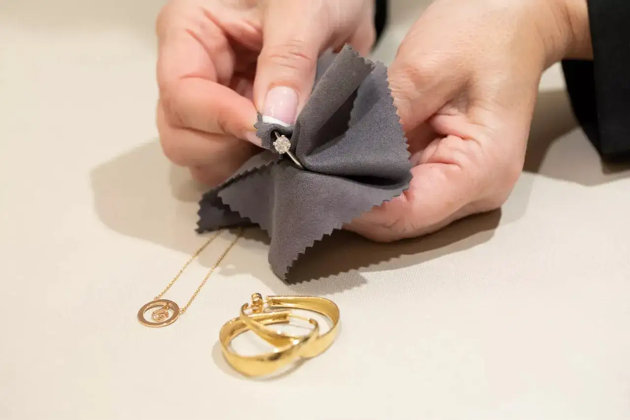 Manos limpian un anillo de oro con un paño gris. Un collar y unos pendientes de oro yacen cerca. Evita que el oro se ponga negro.