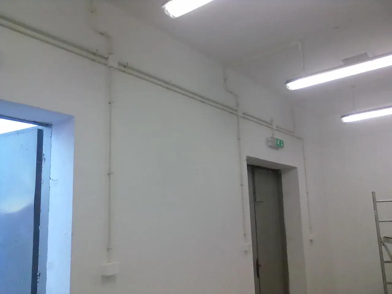 Białe rury tworzą instalację elektryczną natynkową na jasnej ścianie. Widać drzwi i oświetlenie.