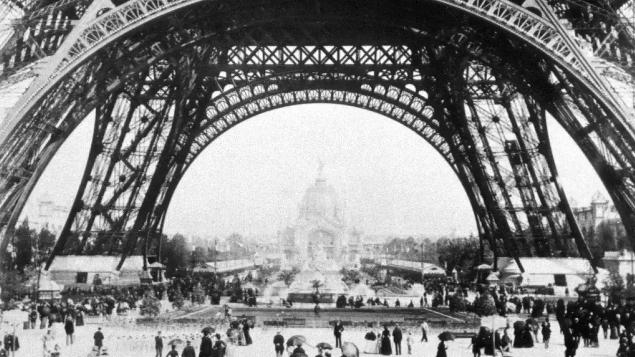 Blick unter den Eiffelturm, der die **Eiffelturm Geschichte** mit Menschenmassen und Pavillons auf der Weltausstellung 1889 zeigt.
