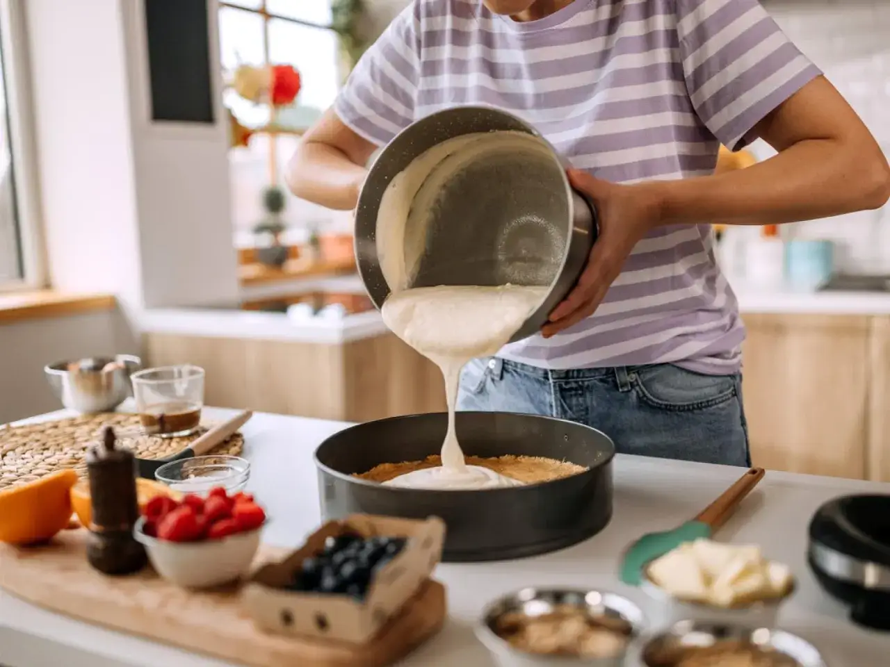 Eine Person gießt eine cremige Masse in eine Springform, um einen Kuchen zu backen. Zutaten wie Beeren und Orangen liegen bereit.