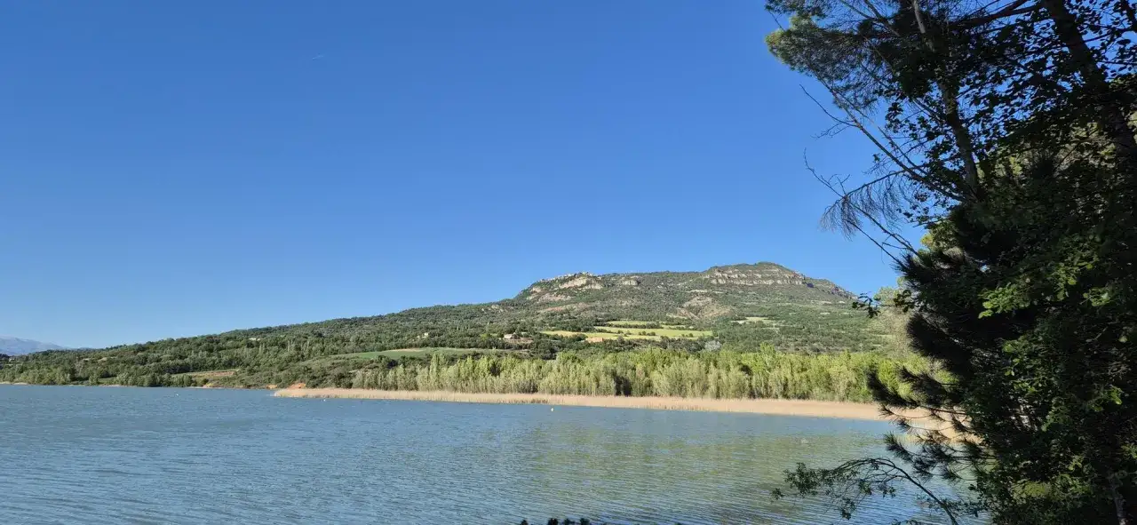 El estany de Basturs, con su agua serena y el verde de la vegetaci&oacute;n, se extiende bajo un cielo azul intenso.