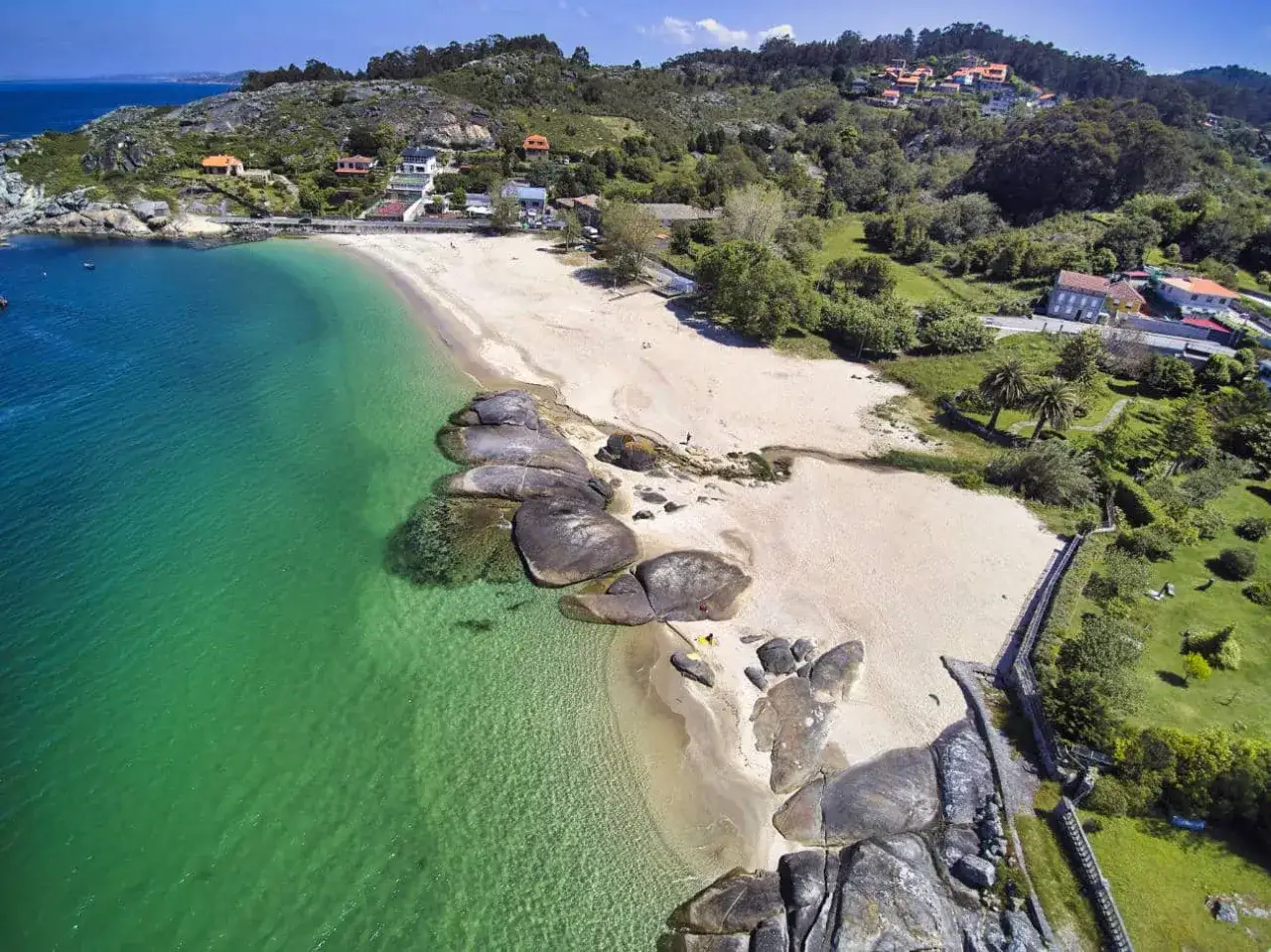 Vista aérea de la **playa pintens**, con arena dorada, aguas cristalinas y rocas. Casas y vegetación verde cubren las colinas circundantes.