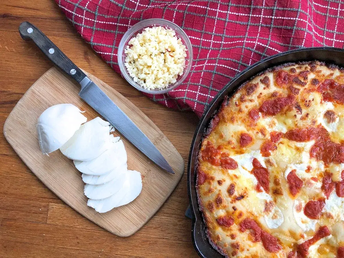 Gotowy ser do pizzy, pokrojona mozzarella na desce, nóż i miseczka z posiekanym czosnkiem.