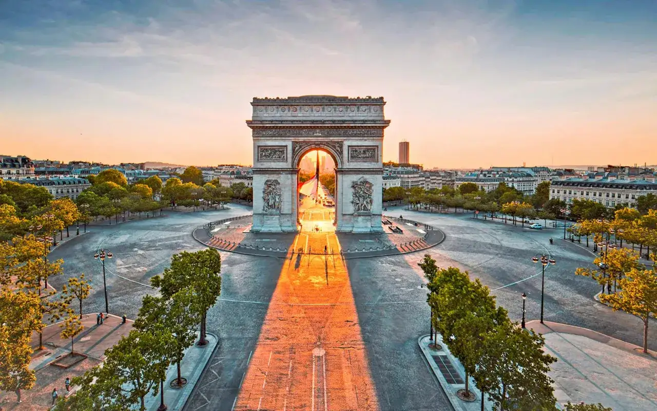 Parisisk morgonljus str&aring;lar genom triumfb&aring;gen. En ikonisk vy av Paris, med dess storslagna arkitektur och gr&ouml;na tr&auml;d.