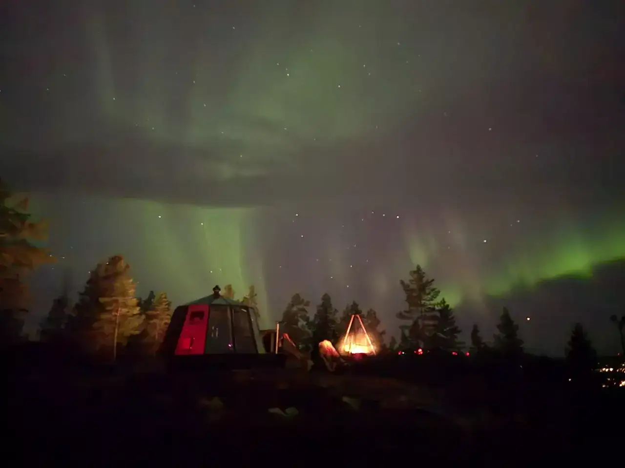 Glamping i Norrland under norrskenet. En glaskupol och ett t&auml;lt lyser upp natten medan himlen dansar i gr&ouml;nt.