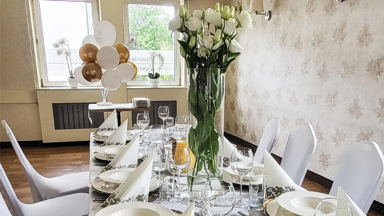 Elegancko nakryty st&oacute;ł w restauracji, ozdobiony białymi kwiatami i balonami. Idealne miejsce na uroczystość, jak chrzciny, kt&oacute;re mogą trwać wiele godzin.