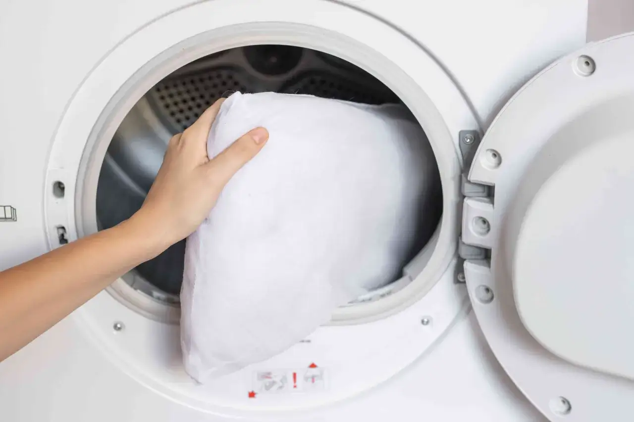 A hand places a white pillow into a washing machine, demonstrating how to wash pillows for a fresh night's sleep.