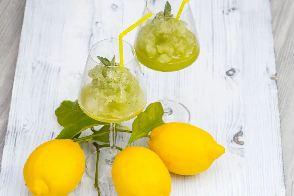 Due bicchieri di sorbetto al limone senza albume, guarniti con menta e cannucce gialle, accanto a limoni freschi su un tavolo di legno bianco.