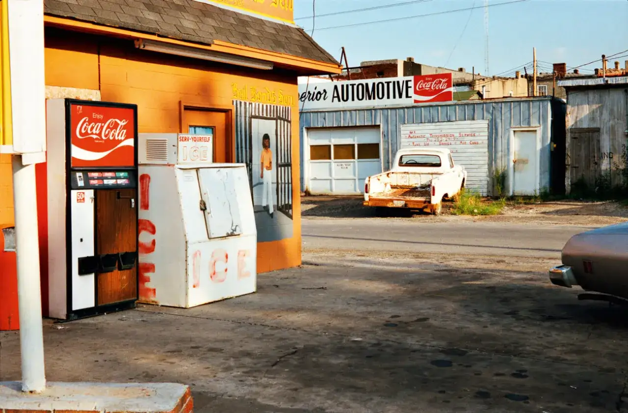 William Eggleston foto: distributore Coca-Cola e un vecchio pick-up bianco davanti a un'officina, con un'atmosfera tipica americana.