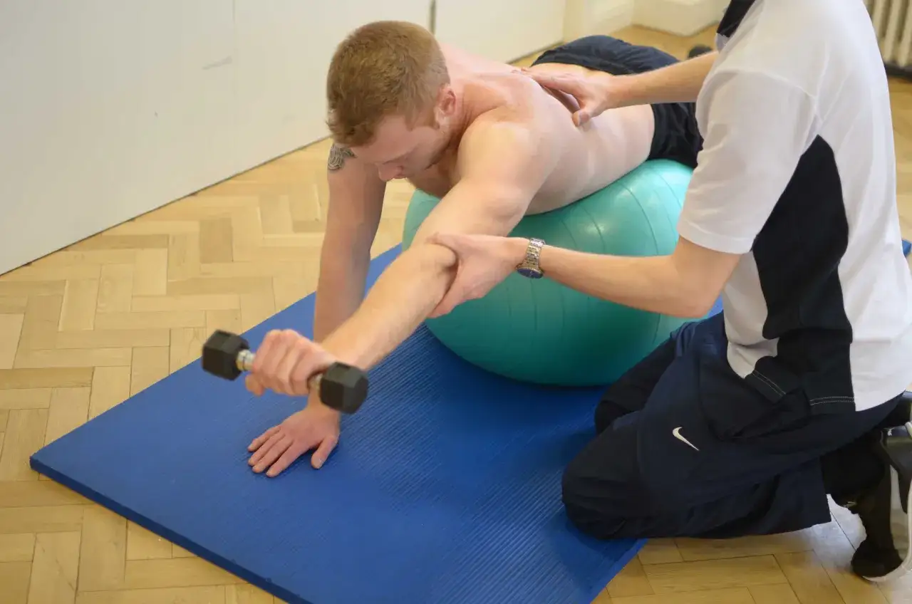 Mężczyzna z chorobą Scheuermanna ćwiczy z hantlem na piłce gimnastycznej, rehabilitant pomaga mu w ćwiczeniach.