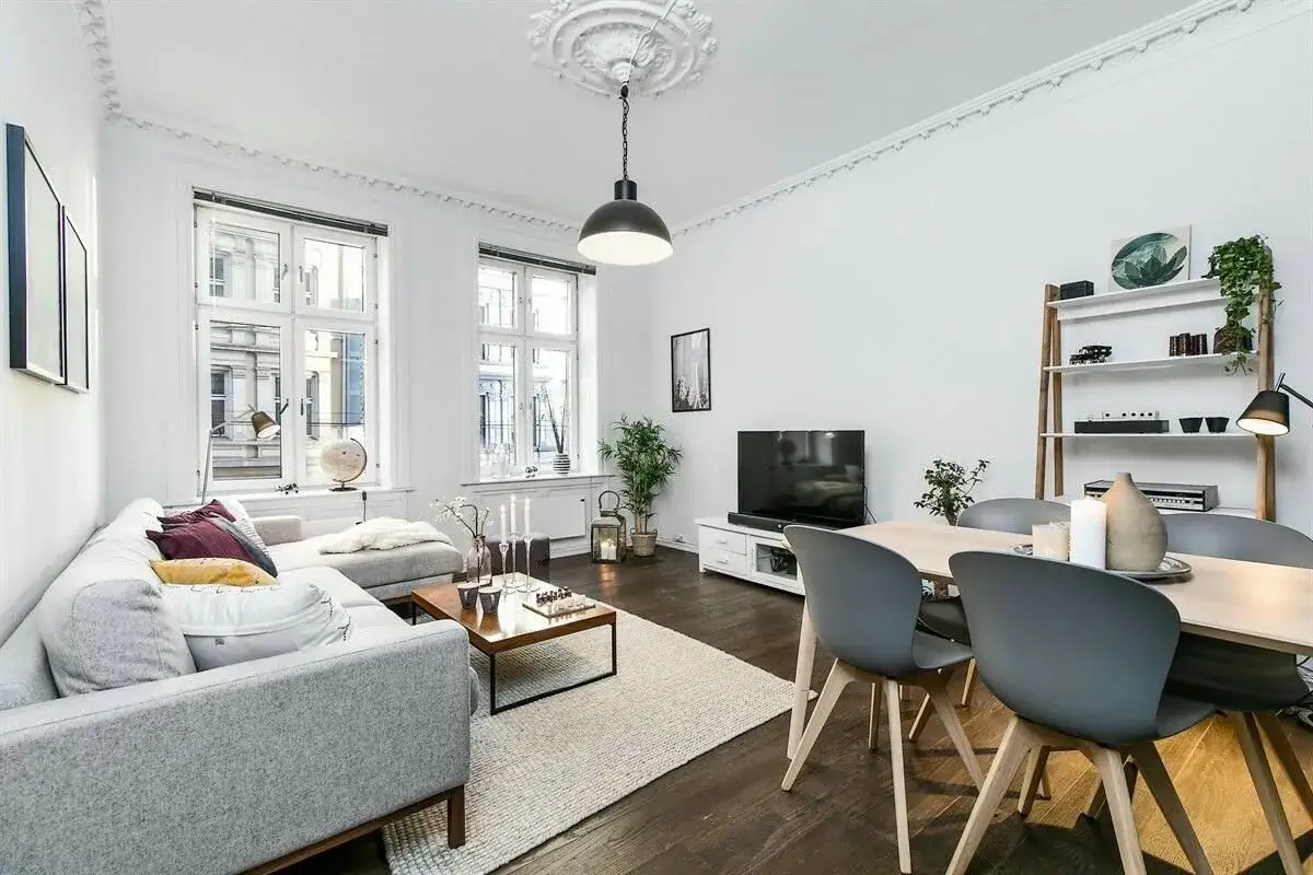 Elegante salón con sofá gris, mesa de centro y comedor. El **suelo oscuro** de madera contrasta con las paredes blancas y la luz natural.