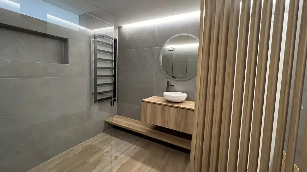 Modernos **ideas ba&ntilde;os**: lavabo sobre mueble de madera, espejo redondo, ducha con toallero negro y panel de lamas de madera.