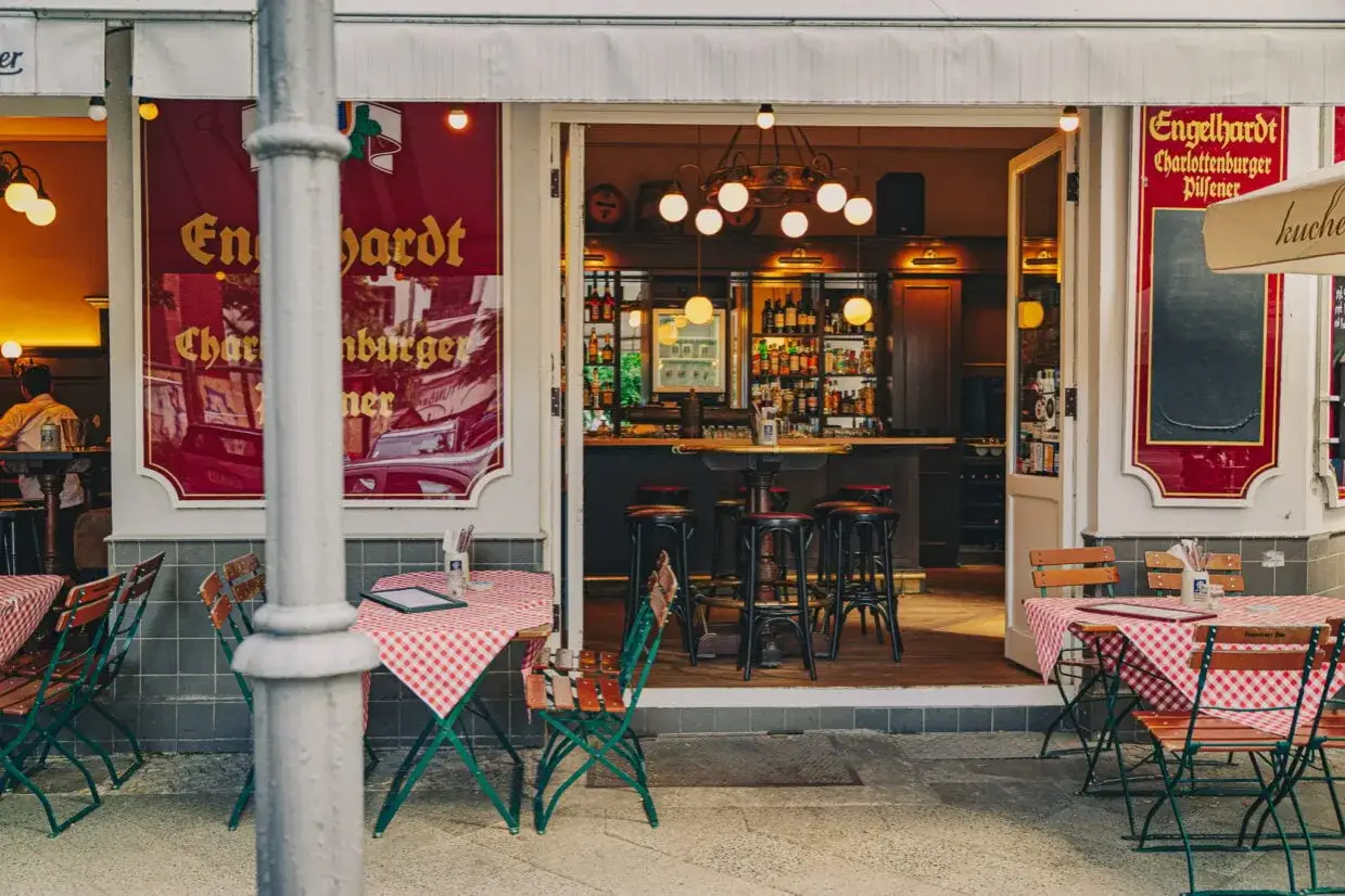 Gemütliche Biergarten-Atmosphäre vor einem Restaurant mit roten karierten Tischen. Ein Schild wirbt für
