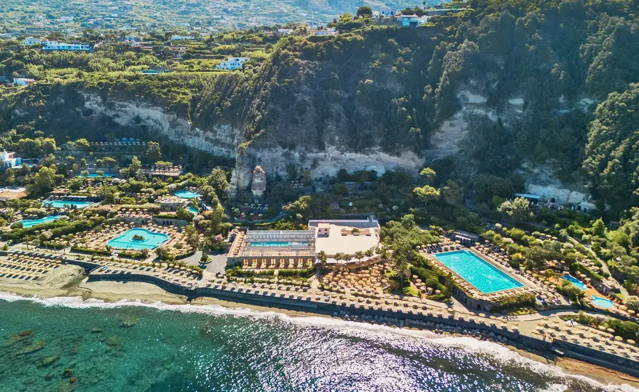Vista aerea di un resort con piscine e lettini sulla spiaggia, incastonato tra il mare e una scogliera verdeggiante. Un paradiso per le terme a Forio.