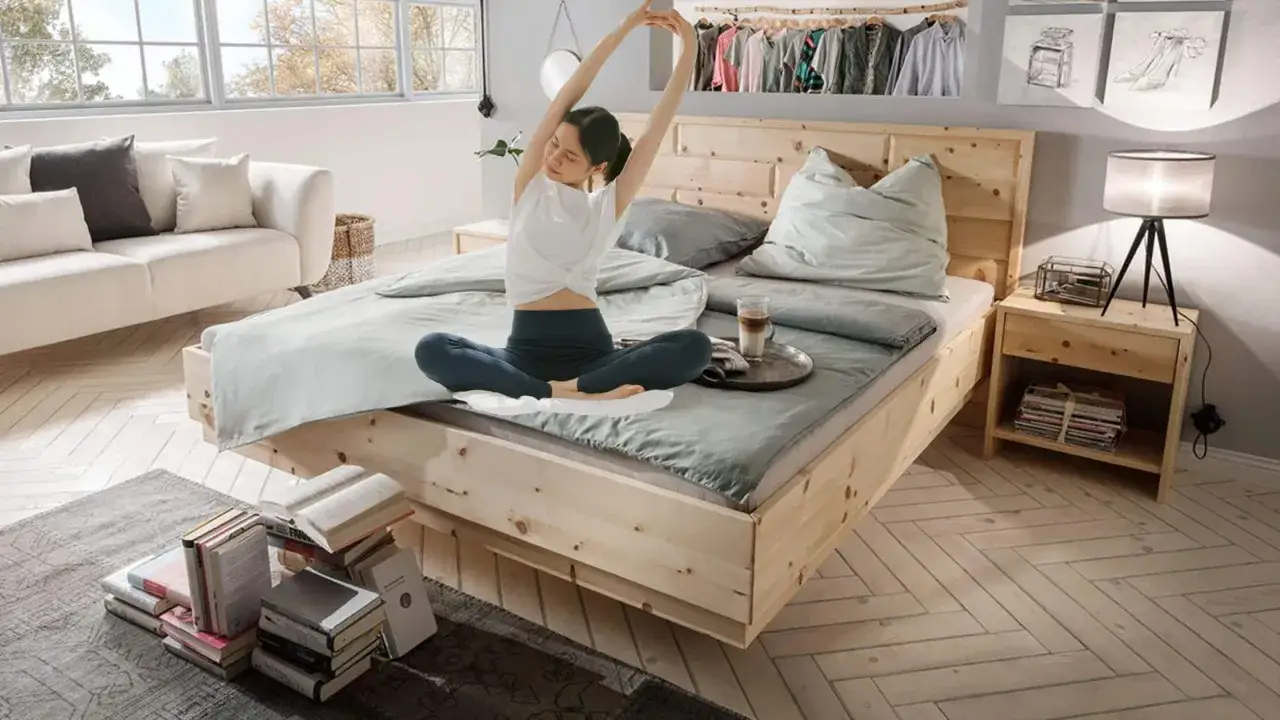 Frau dehnt sich im hellen Schlafzimmer, das mit einem Holzbett und Sofa eingerichtet ist. Ein Nachttisch mit Lampe und Büchern vervollständigt das Bild.