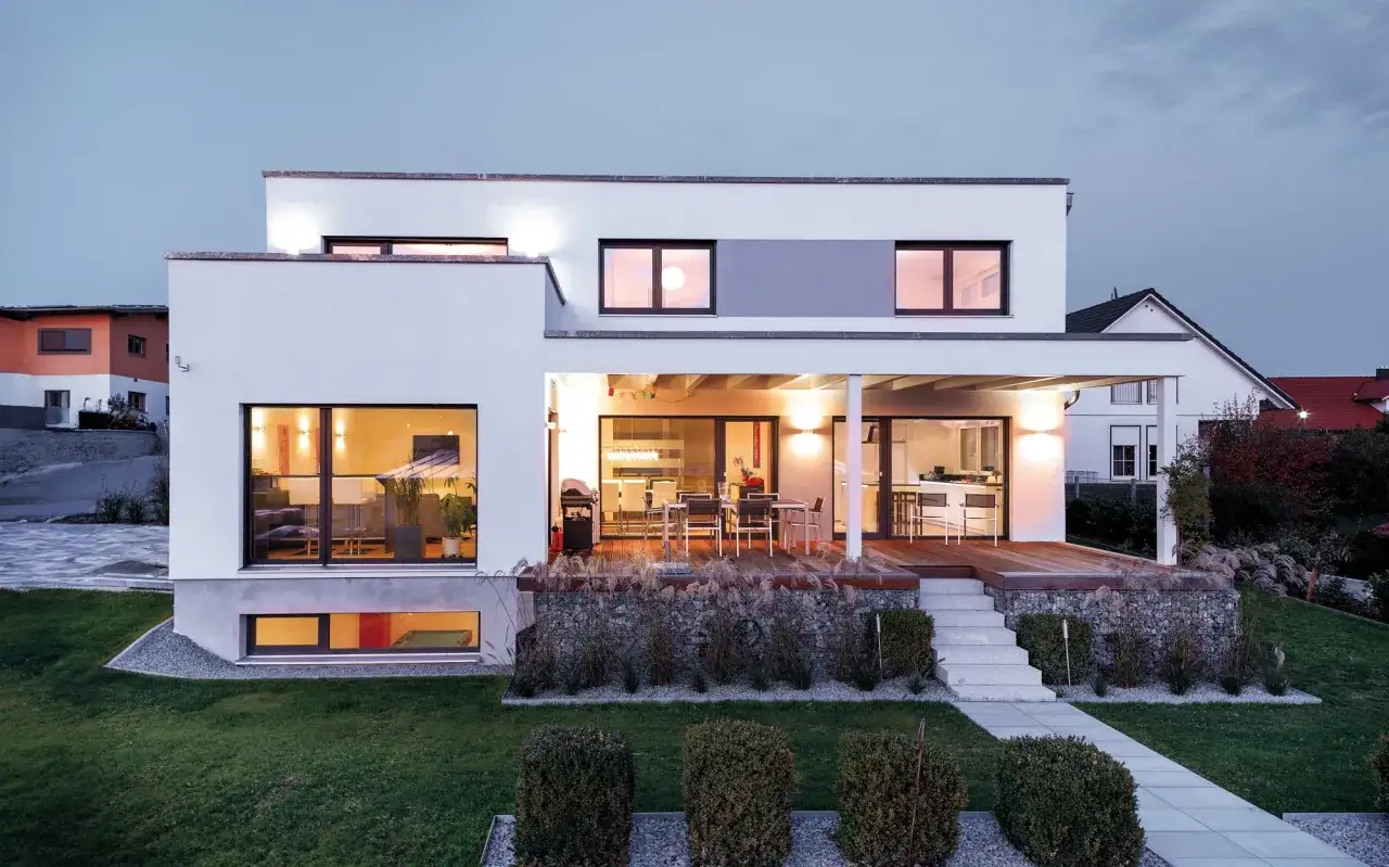 Moderner Fertighaus Kubus mit Terrasse und großzügigen Fensterfronten, beleuchtet im Abendlicht.