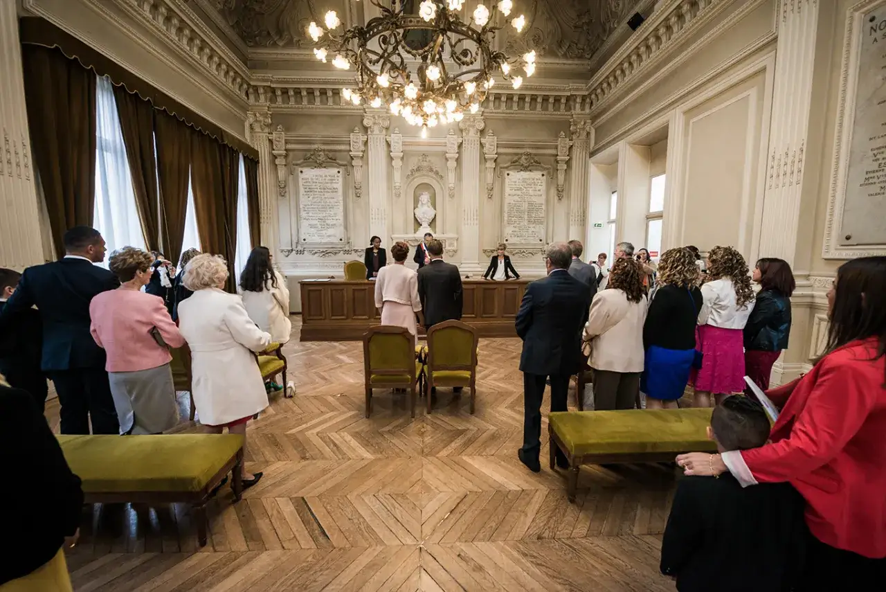 Une cérémonie de mariage se déroule dans une salle solennelle, avec des invités observant le couple devant l'officier.