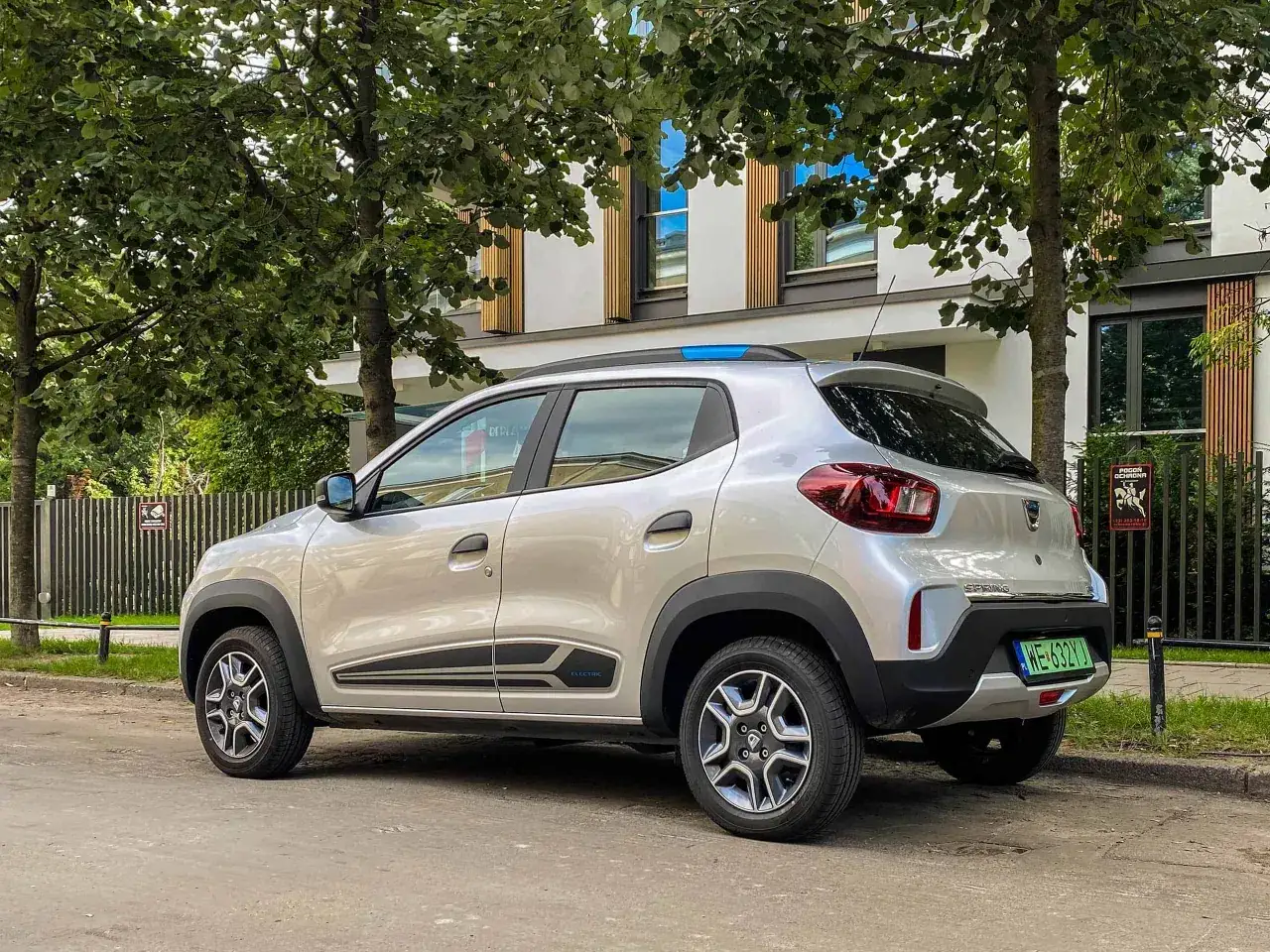 Srebrna Dacia Spring, elektryczny samochód miejski, zaparkowany pod drzewami. Długi dacia spring zasięg pozwala na swobodne podróżowanie po mieście.