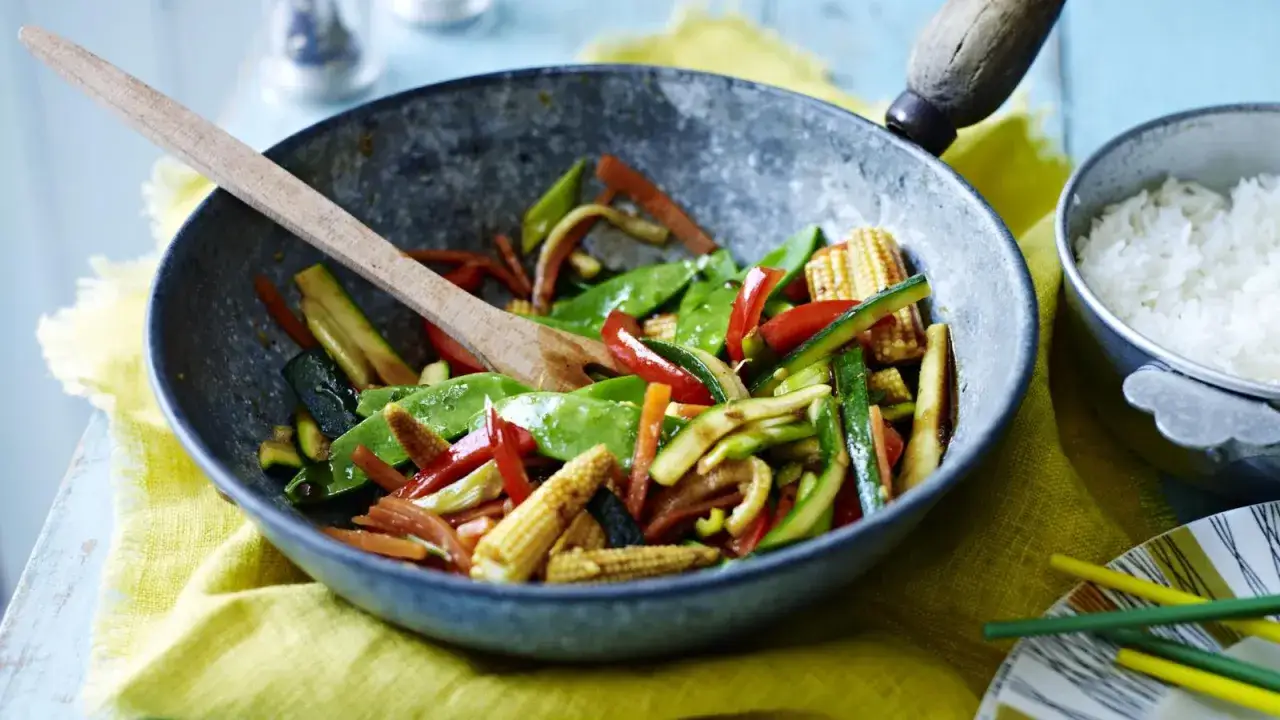 Kolorowy stir fry z cukinią, papryką, groszkiem cukrowym i kukurydzą, mieszany drewnianą łyżką w woku. Obok miseczka ryżu.