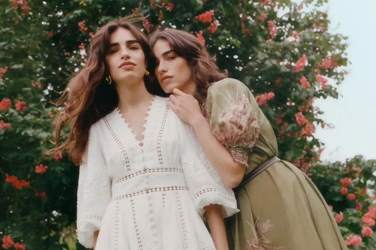Two women embrace in a garden, embodying the spring chic dress code. One wears a white lace dress, the other a floral green gown.