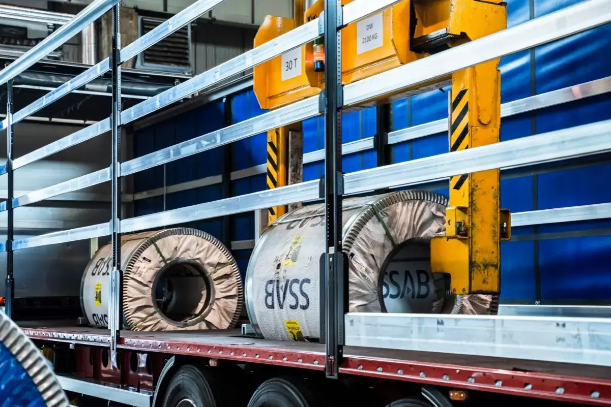 Ciężarówka z wielkimi, metalowymi kręgami coilmulda, gotowa do transportu. Żółty dźwig czeka na załadunek.