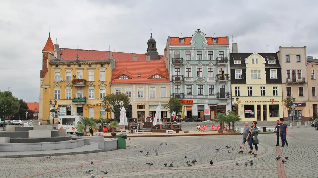 Kolorowe kamienice zdobią rynek w Gnieźnie. Na bruku spacerują ludzie, a gołębie szukają jedzenia.