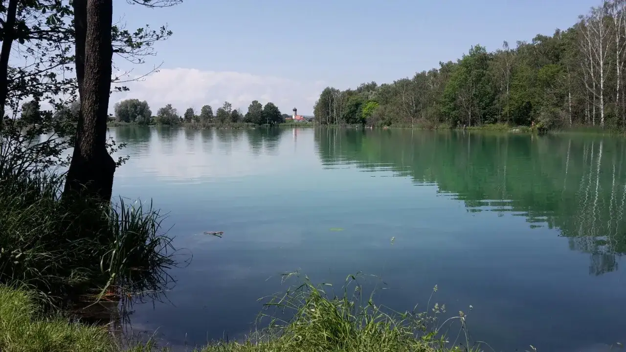 Idyllischer ersinger badesee mit gr&uuml;nen Ufern und einem Kirchturm in der Ferne.