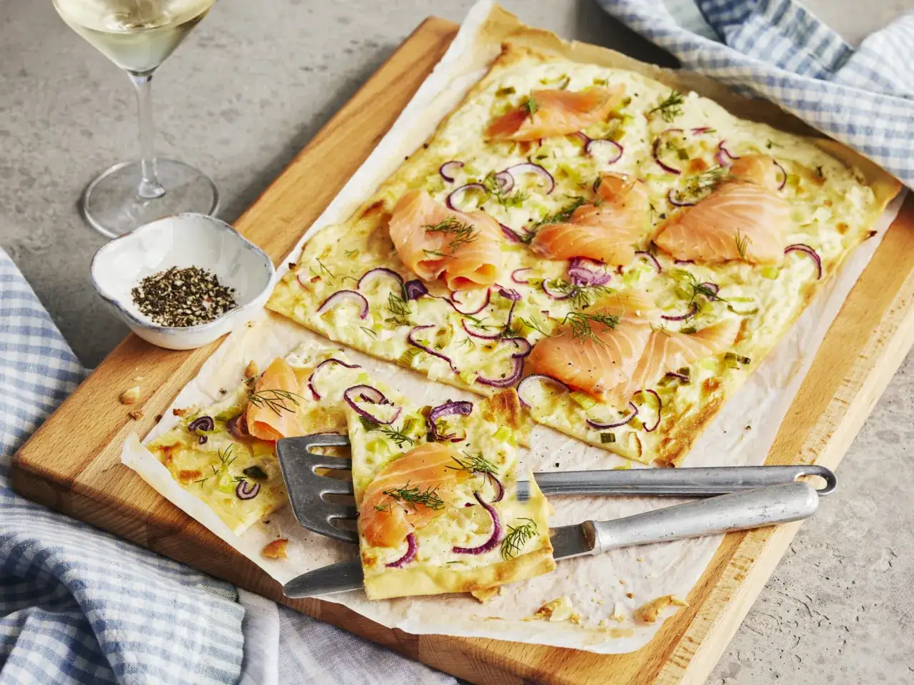 Flammkuchen mit Lachs, Zwiebeln und Dill auf einem Holzbrett. Ein Glas Wei&szlig;wein und eine Schale Pfeffer stehen daneben.