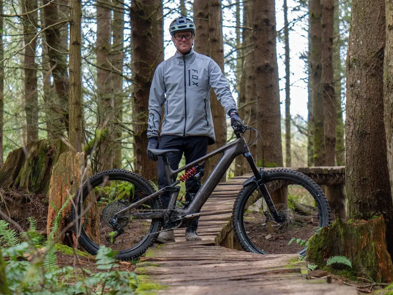 A man in a grey FOX jacket and helmet stands with his specialized Levo 4 mountain bike on a wooden trail in a forest.