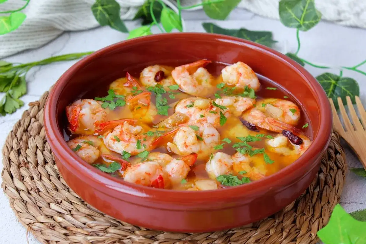 Langostinos al ajillo burbujeantes en cazuela de barro, con perejil fresco picado y un toque de guindilla.