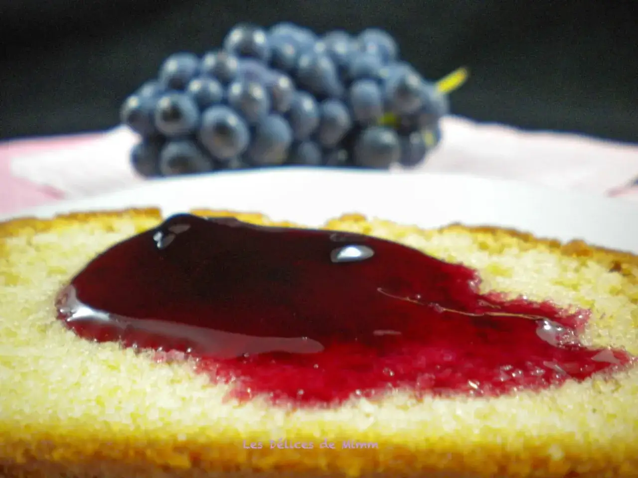 Tranche de gâteau moelleux nappée d'une généreuse gelée de raisin, avec une grappe de raisins bleus floue en arrière-plan.