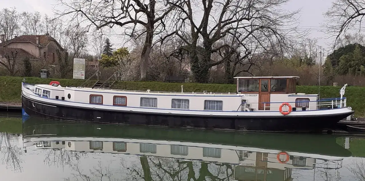 Une p&eacute;niche blanche et noire, reflet d'une vie paisible sur l'eau, est amarr&eacute;e pr&egrave;s d'une maison ancienne.