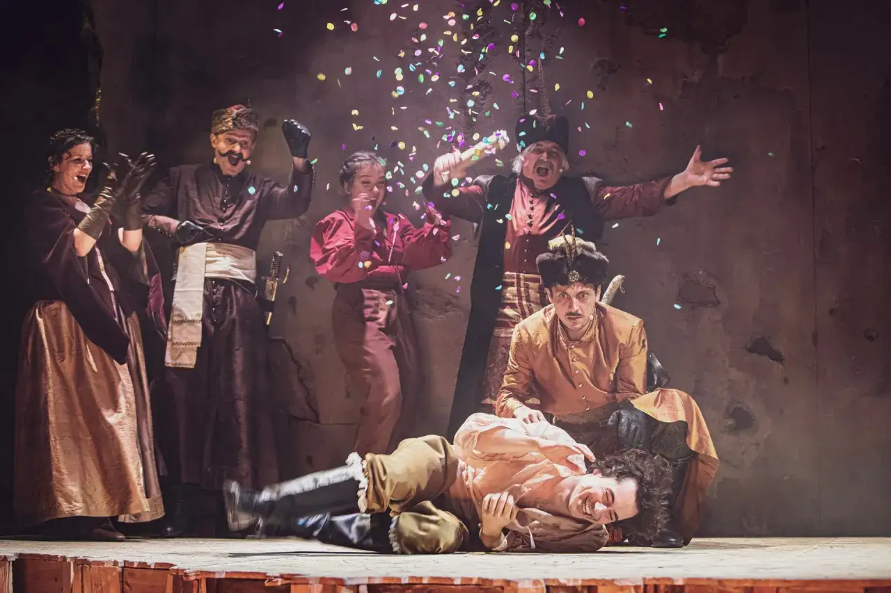 Aktorzy w strojach ludowych świętują na scenie, sypiąc konfetti. To fragment repertuaru teatru ludowego.