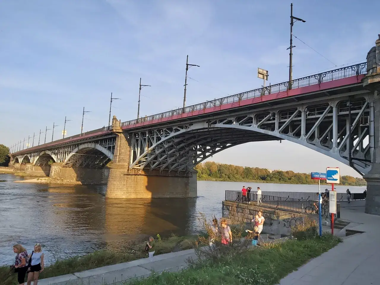 Most Poniatowskiego w Warszawie, z charakterystyczną stalową konstrukcją i czerwonymi elementami, nad spokojną Wisłą.