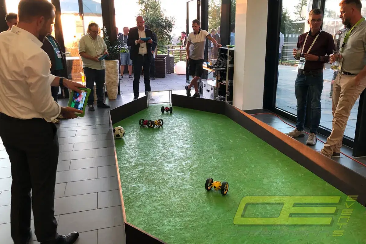 Männer steuern ferngesteuerte Autos auf einem grünen Spielfeld, ein Fußball liegt daneben. Ein spannender **Drohnen Sport**-Moment.