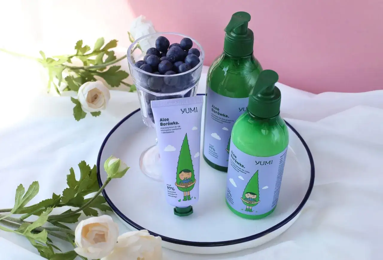 Krem do rąk i balsam do ciała YUMI Aloe Borówka, z uroczą grafiką, obok szklanki z borówkami i kwiatów.