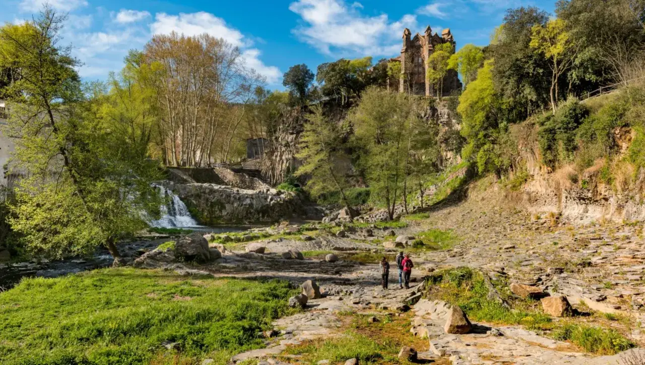 Senderismo en el parc natural de la zona volc&agrave;nica de la Garrotxa, con un castillo en ruinas sobre un acantilado y una cascada.