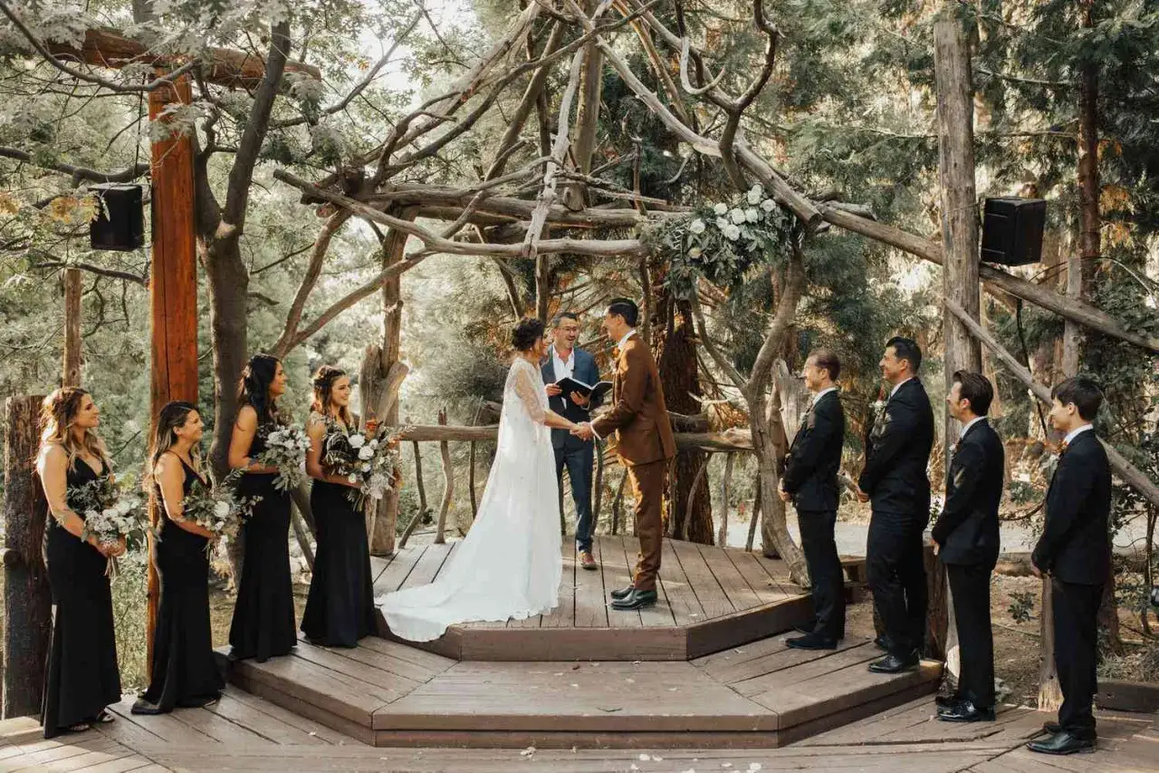 Ein Brautpaar gibt sich das Ja-Wort bei einer romantischen **Hochzeit im Wald**. Die Zeremonie findet unter einem kunstvollen Holzpavillon statt, umgeben von Bäumen.