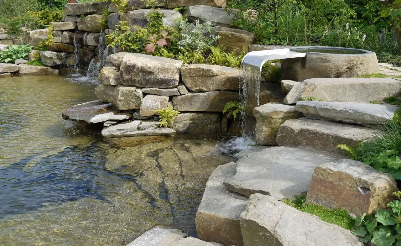 Ein moderner Wasserfall aus Stein und Metall, der zum **wasserfall selber bauen** inspiriert. Das Wasser plätschert in einen Teich.