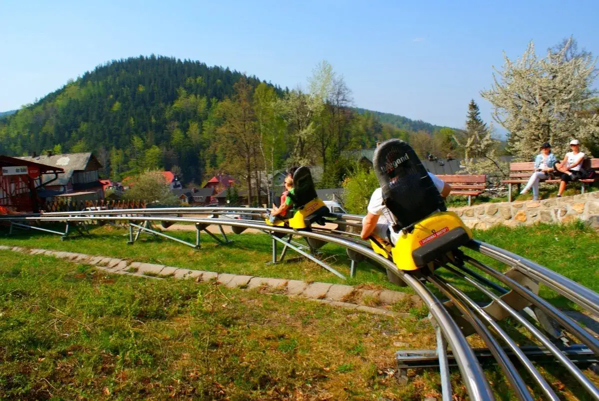 Tor saneczkowy w **centrum rekreacji i sportu kolorowa** w g&oacute;rach. Dwie osoby zjeżdżają na sankach, w tle zielone wzg&oacute;rza i domki.