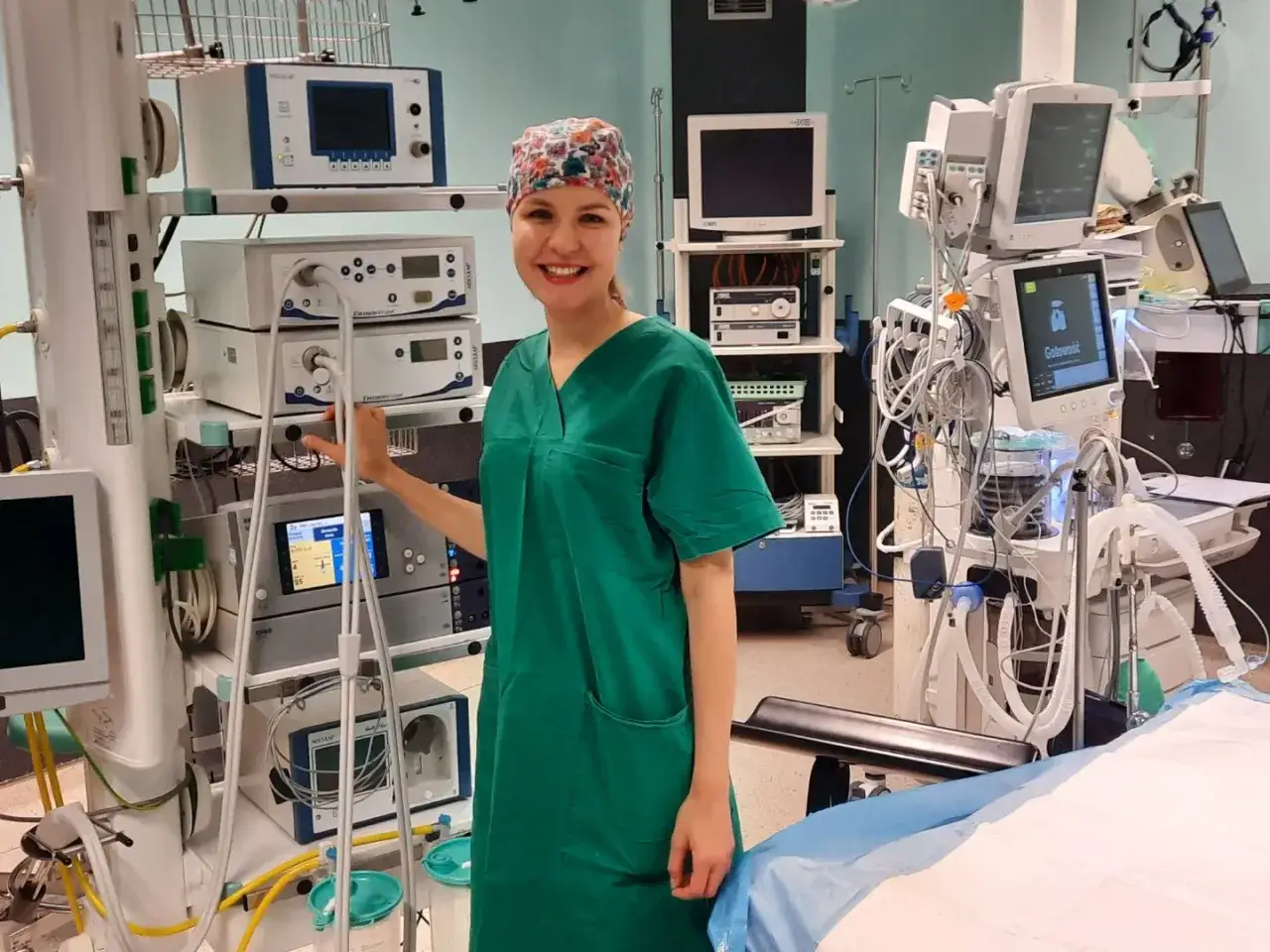 Uśmiechnięta pielęgniarka operacyjna, w zielonym fartuchu i czepku, stoi obok aparatury medycznej. Obok niej widać instrumentariuszka przygotowująca się do zabiegu.