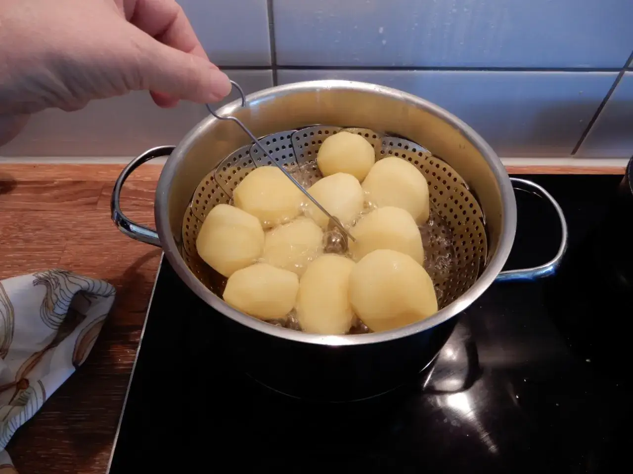 En hand lyfter en sil med skalade potatisar ur en kastrull med bubblande vatten. Perfekt för att koka potatis med eller utan lock.