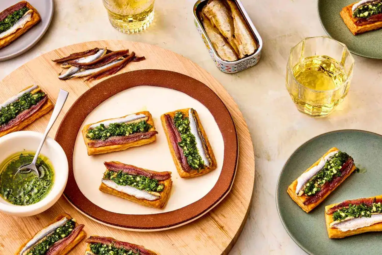 Rectangular pastries topped with a vibrant green herb mixture and delicate white anchovies, served with a side of oil and a tin of preserved fish.