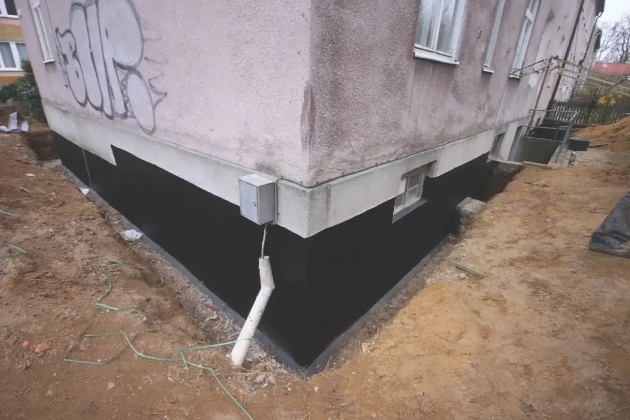 Prace przy osuszaniu fundamentów budynku. Czarna izolacja przeciwwilgociowa na ścianie fundamentowej, wykop z ziemią i rurą odpływową.