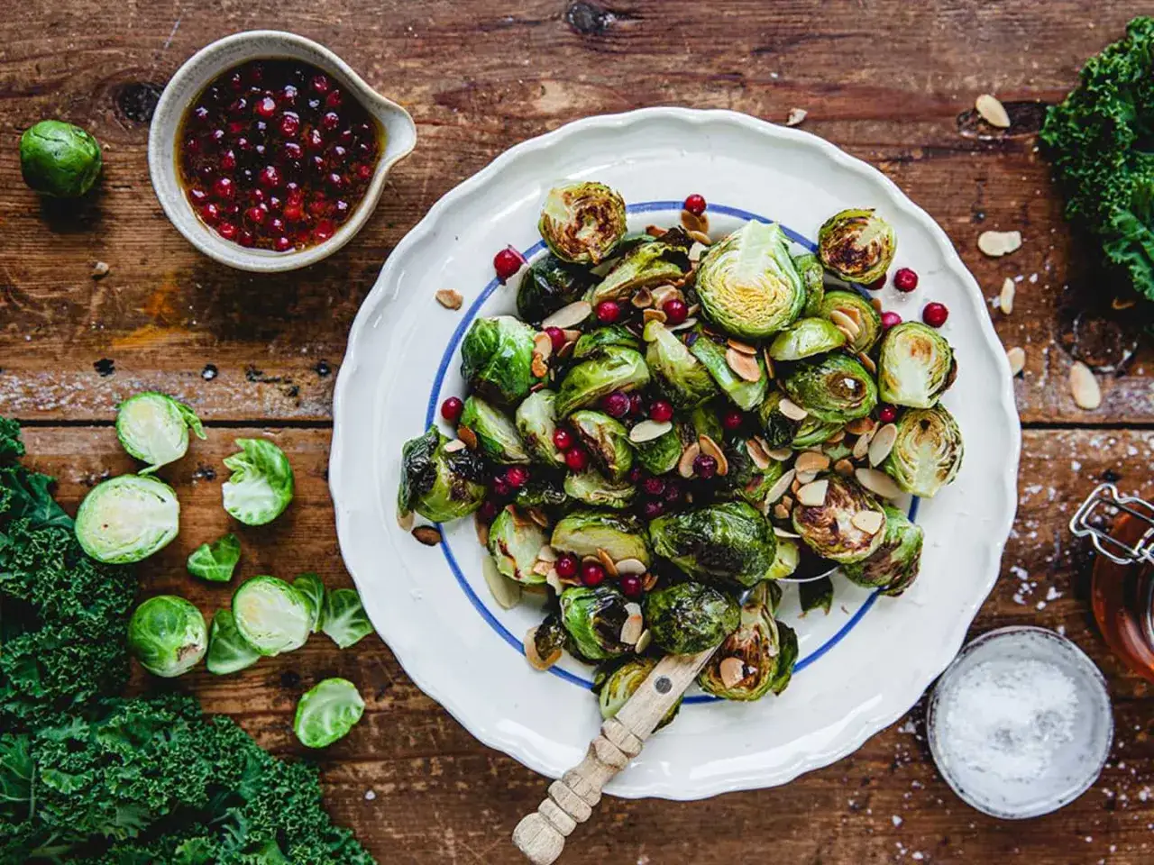 Rostade coles de bruselas med tranb&auml;r och mandel, serverade p&aring; en vit tallrik med bl&aring; kant. En sk&aring;l med dressing och f&auml;rsk gr&ouml;nk&aring;l ligger bredvid.