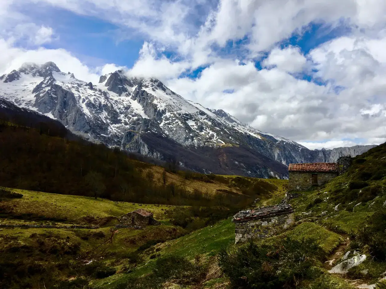 Casas de piedra en una ladera verde, con montañas nevadas al fondo. Un camino serpentea, evocando la **ruta de la reconquista**.