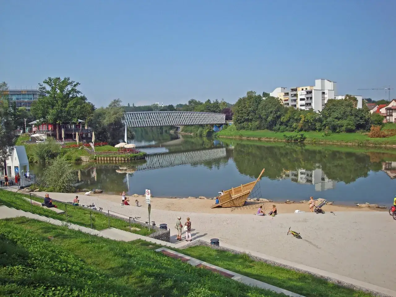 Sonniger Tag am Neckarstrand Remseck mit Menschen am Sandstrand, einem Spielschiff und einer Brücke über dem Fluss.