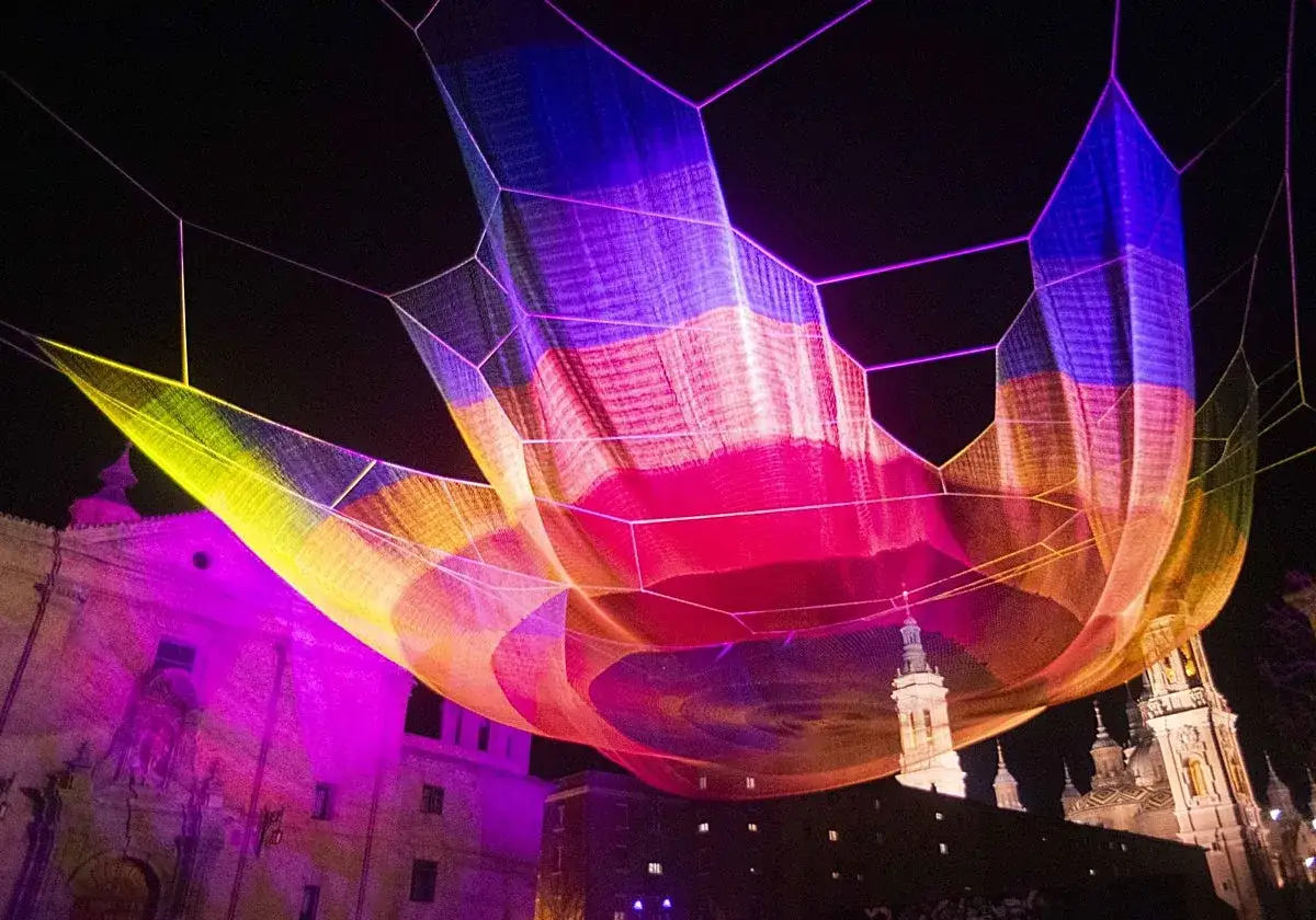 Instalación de luces de colores vibrantes sobre edificios históricos, evocando la magia de una fiesta de los 90 en Zaragoza.