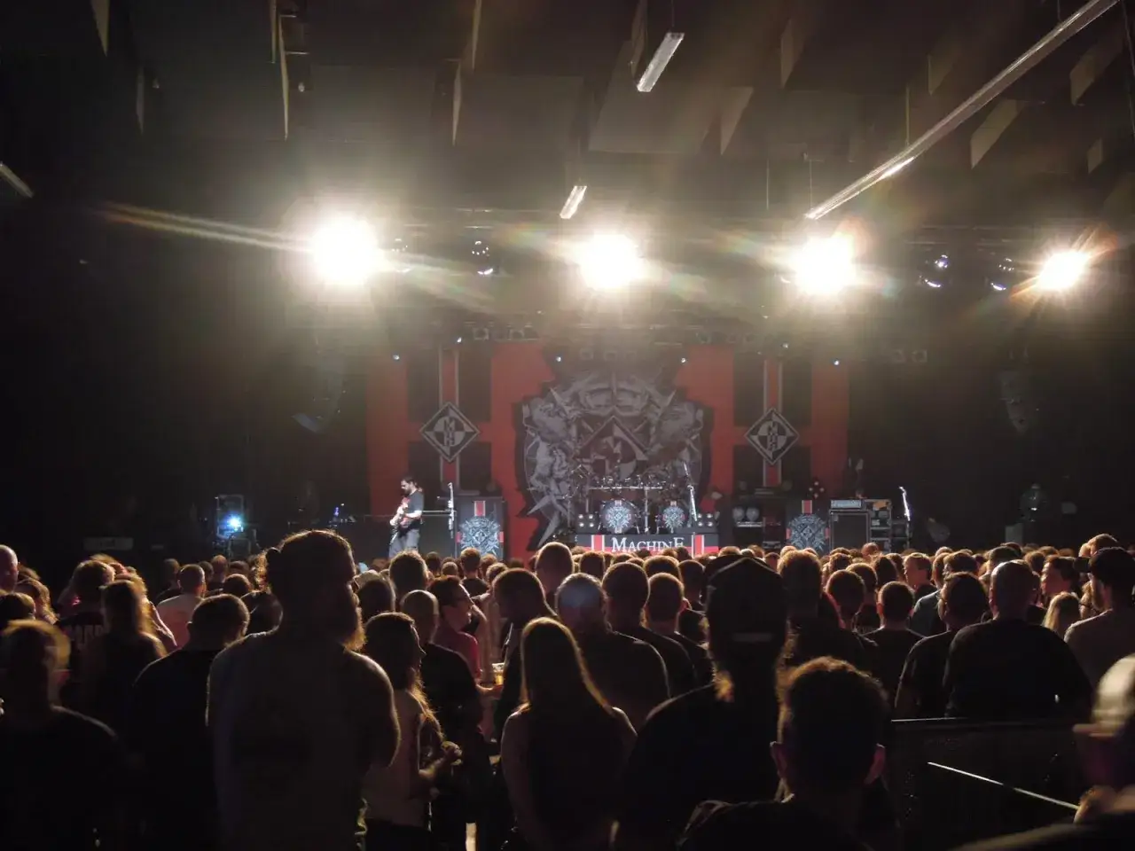 Tłum na koncercie Machine Head, światła sceniczne, muzycy grający na gitarach i perkusji.
